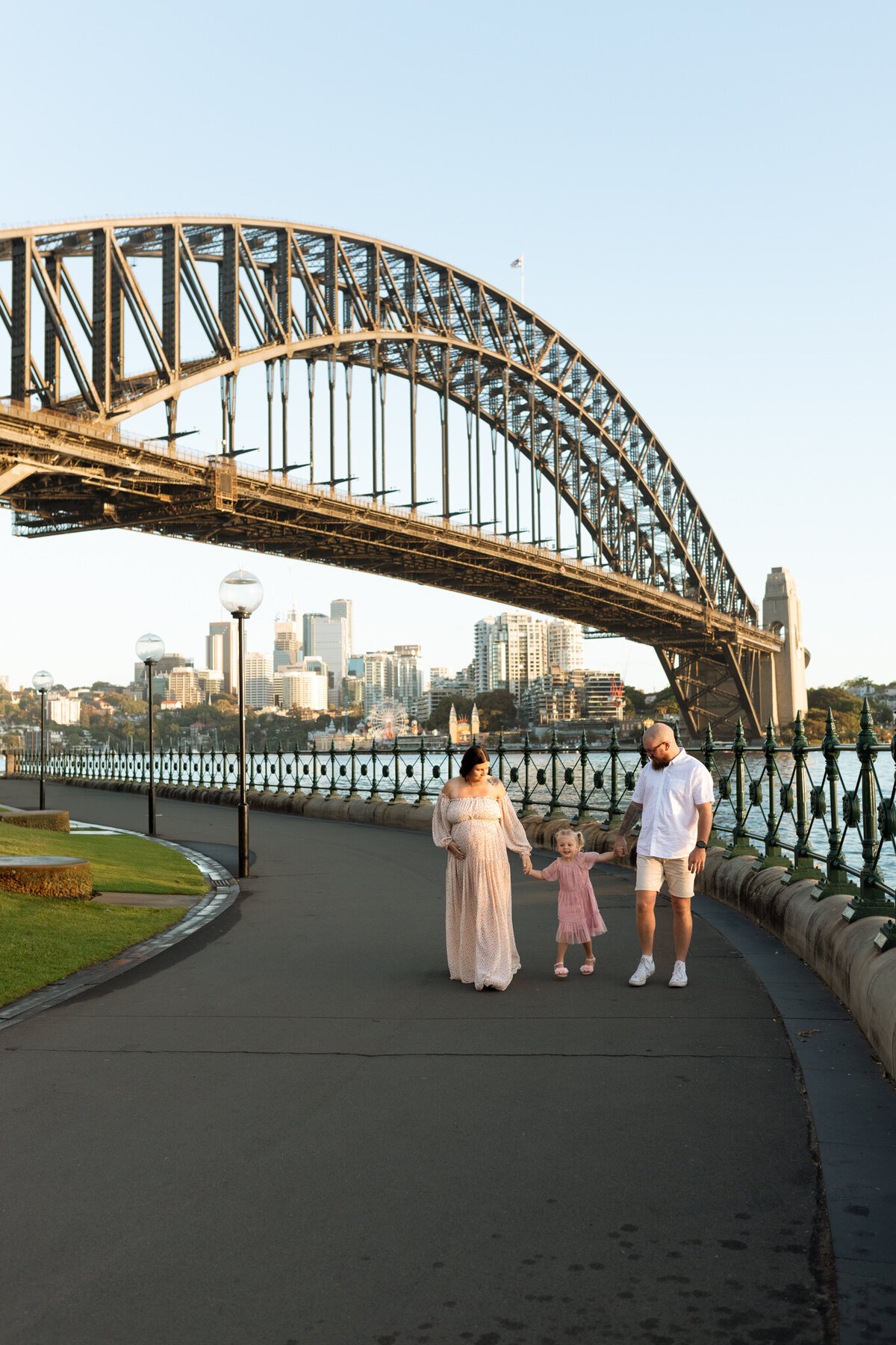 Pregnant woman walking hand in hand with her partner and young daughter beneath the Sydney Harbour Bridge, with the city skyline and water in the background.