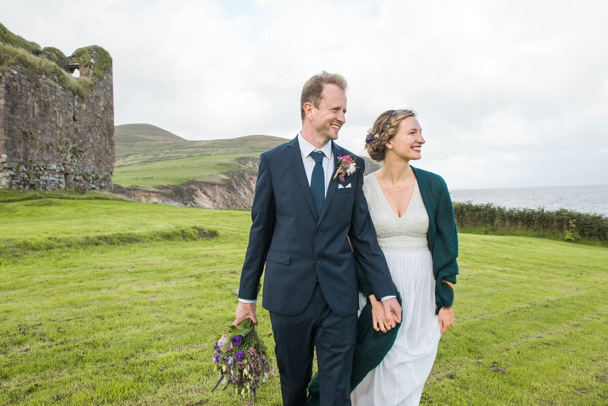 Bride with blonde, vintage hairstyle wearing an empire style wedding dress and draped in a green wool shawl holding hands with her groom wearing a navy suit and tie holding purple wedding bouquet while walking in a field overlooking Minard beach