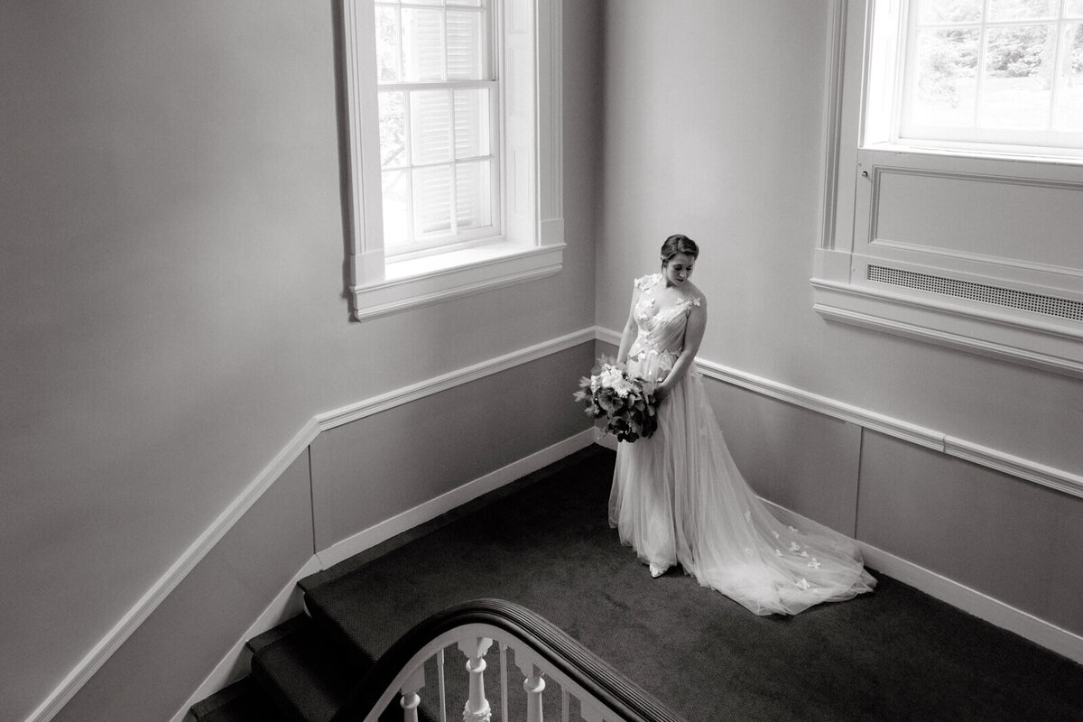 Top view of the bride, holding her flower bouquet, standing on a corner of the house, with large windows in the background.