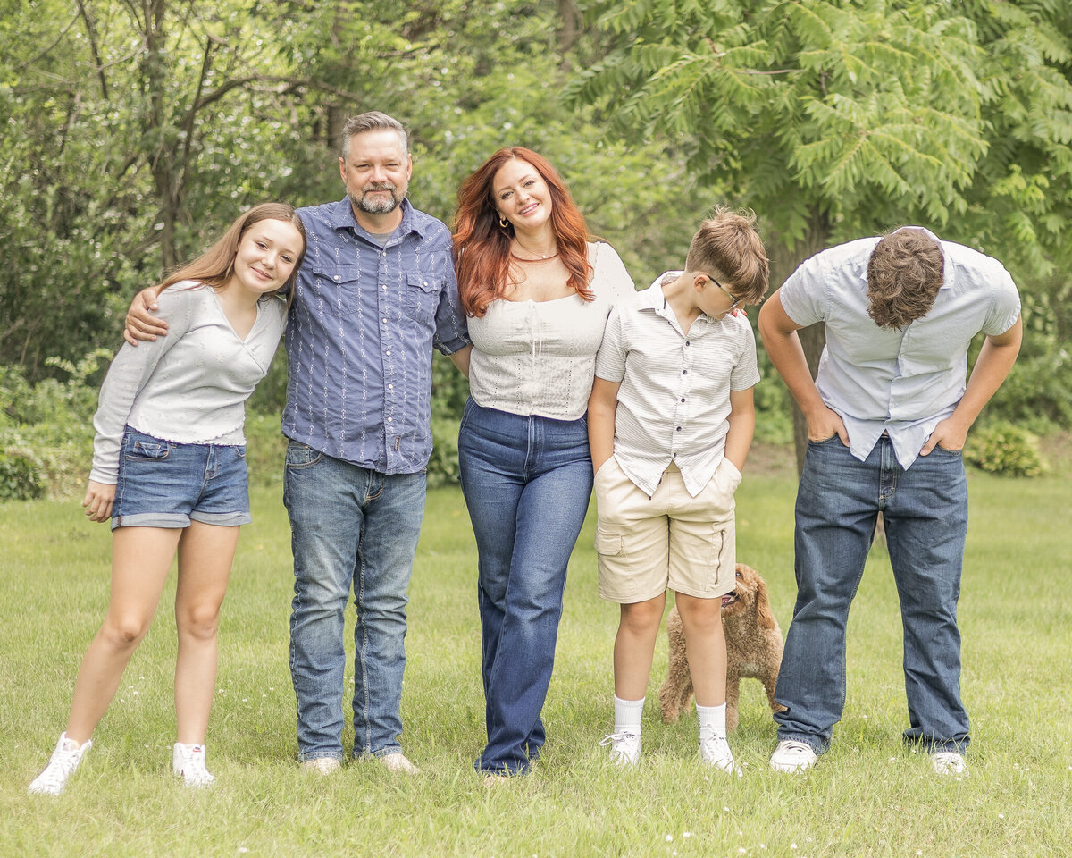 Family of five and their brown dog standing on grass with a tree line behind them.  Mom, Dad, and daughter are looking at the camera smiling while the two brothers are looking down at the dog.Daughter is wearing blue jean shorts, a long sleeve light blue shirt, white shoes, and has long light brown hair.  Dad is wearing blue jeans, blue and white stripped button up shirt, and has short pepper hair and beard.  Mom has long red hair, wearing a white short sleeve shirt and blue jeans.  Younger son is wearing a light tan button up and khaki shorts, white shoes, short light brown hair.  Oldest son is wearing a light blue short sleeve button up, blue jeans, and has short light brown hair