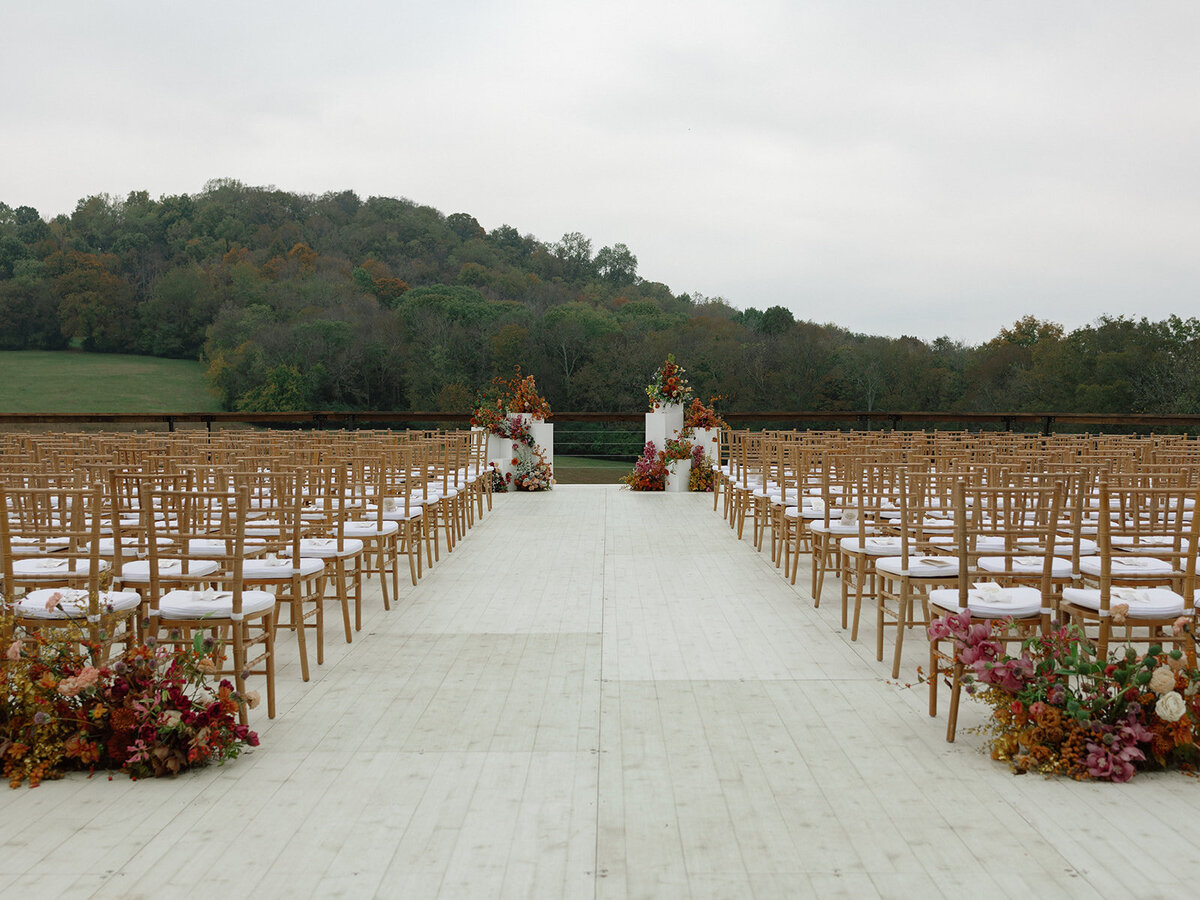 Sculptural columns wedding ceremony backdrop. Stair step white ceremony columns adorned with sculptural floral meadows in fall hues. Clear top tent build out for fall wedding reception in Tennessee hills. Wire threaded orchids, bittersweet berries, butterfly ranunculus, and tulips are highlighted in this wedding ceremony backdrop. Artistic fall color palette in copper, gold, ruby, lavender, and rust. Design by Rosemary & Finch Floral Design in Nashville, TN.