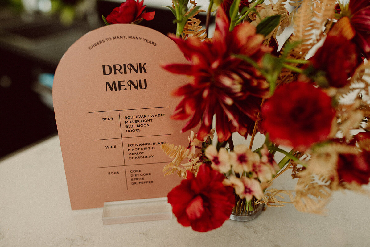 Rounded peach card stock drink menu with black font on clear place card holder atop a white table with wine-colored flowers.