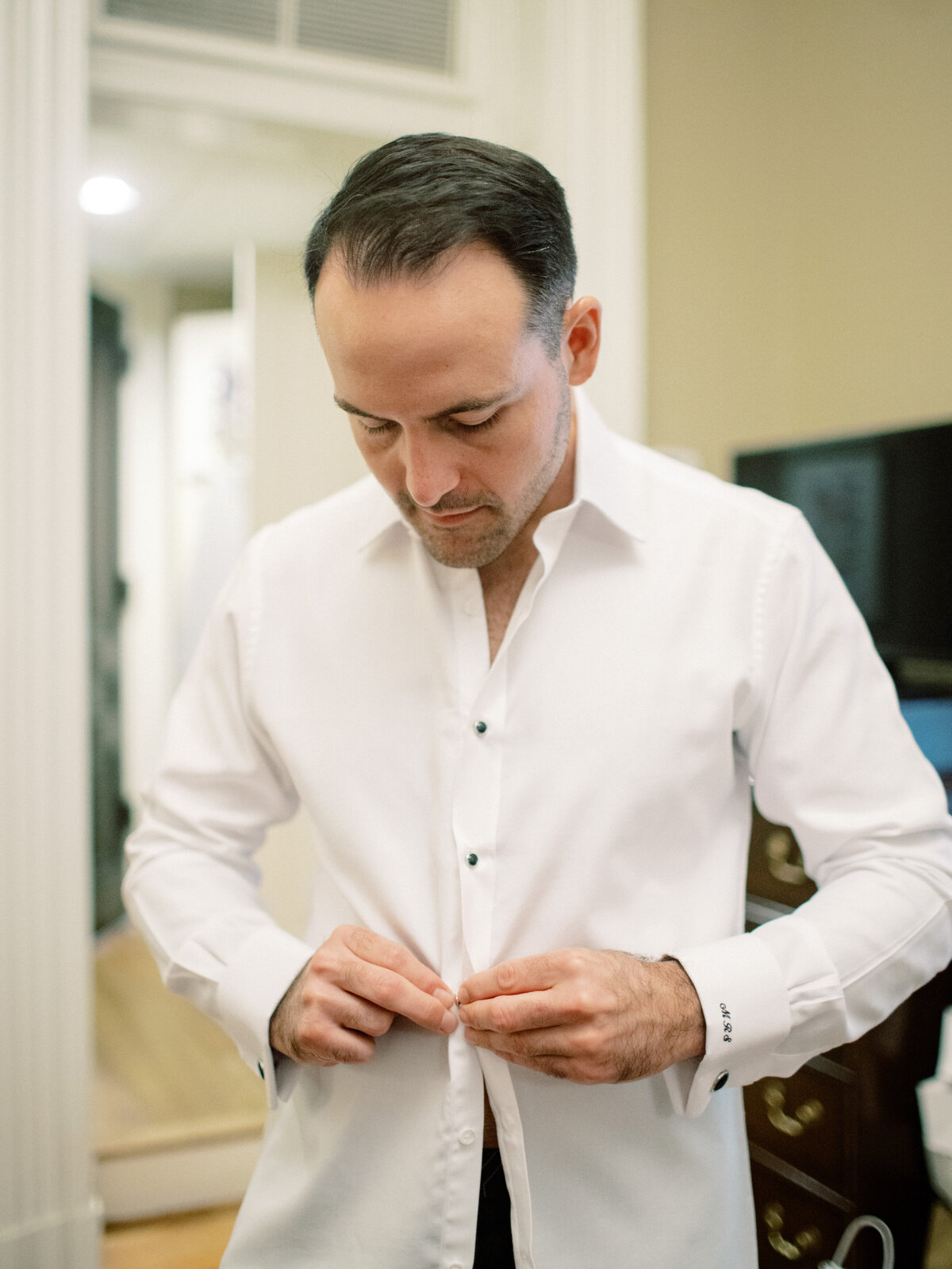 A groom concentrating on buttoning his shirt, highlighting the significance of preparation and attention to detail before the significant day of the wedding.