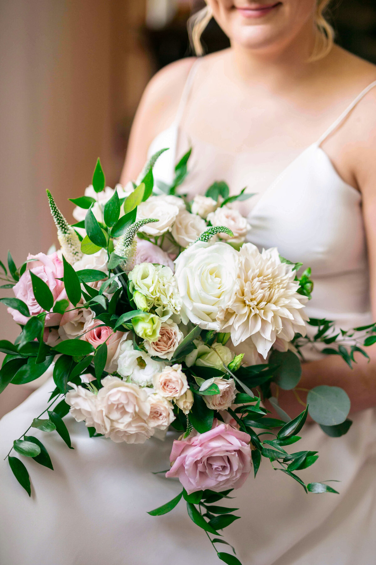 A bride holds a bouquet of white, pink, and green flowers. The bouquet features roses, dahlias, and lush greenery. She is wearing a white gown with thin straps. The focus is on the flowers.