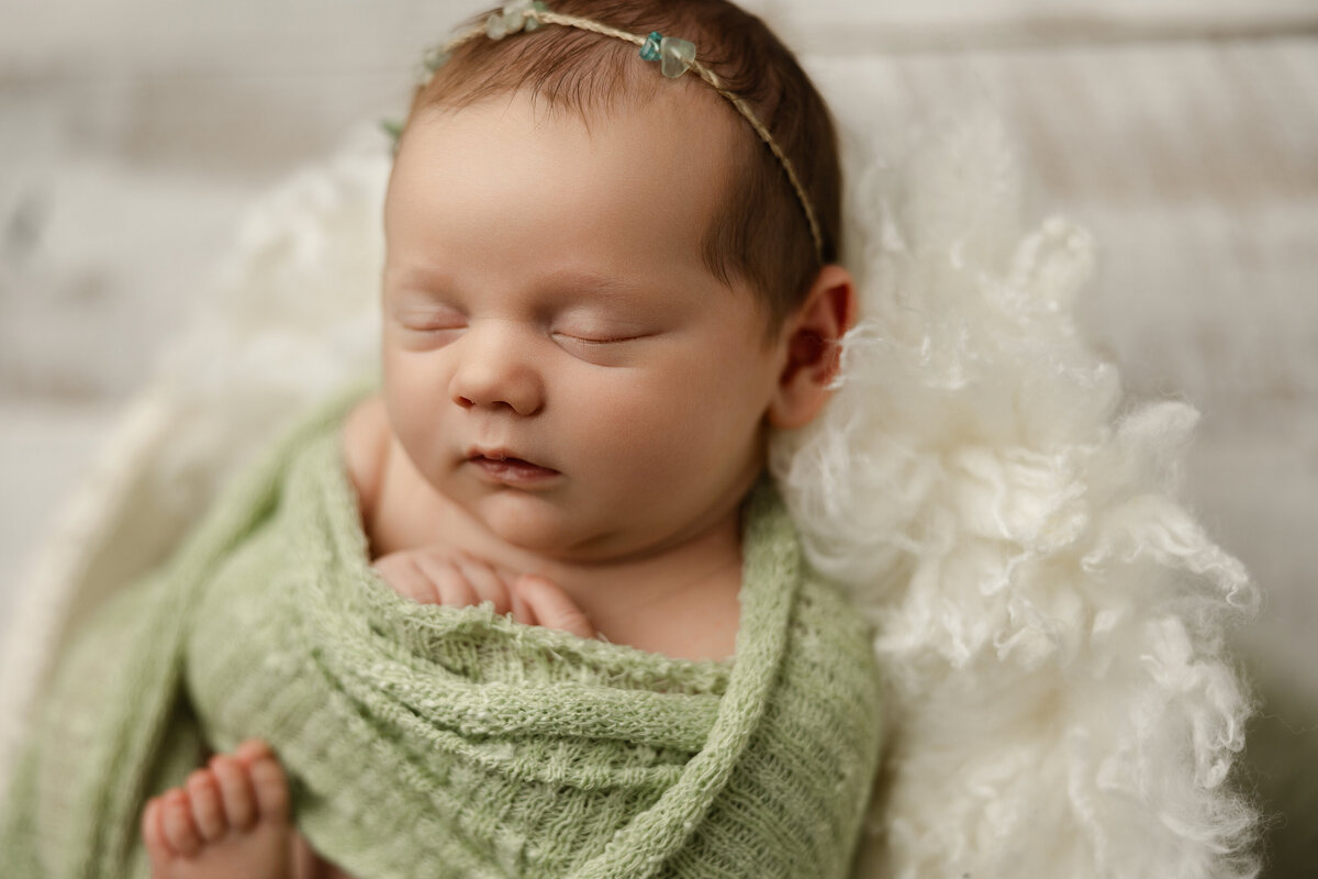 Newborn baby girl wrapped in a light green knit and resting on a fluffy white layer, wearing a delicate floral headband. Captured by Inland Empire newborn photographer Kaitlyn Dawn Photography.