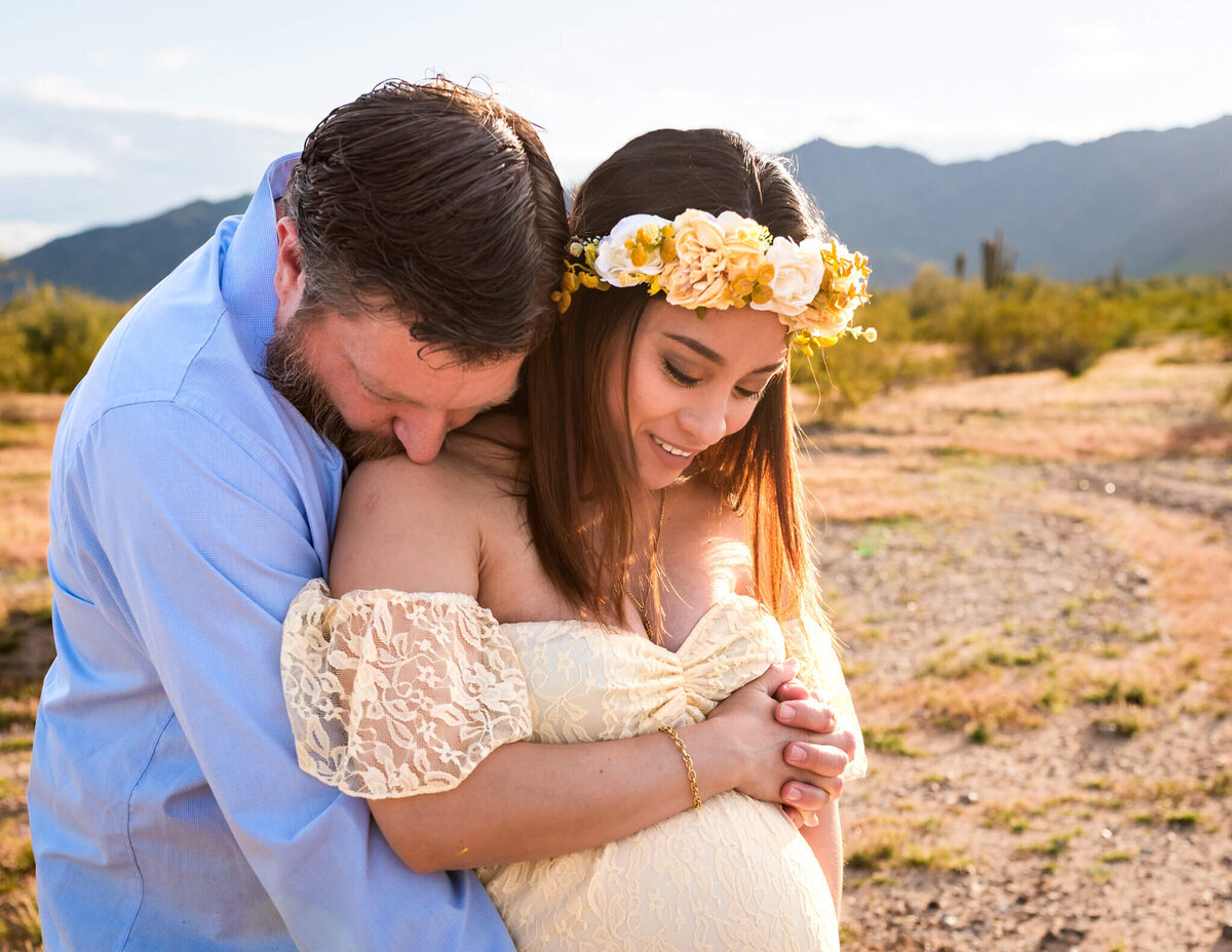 Husband sweetly kissing expecting wife's shoulder while they cradle her baby bump