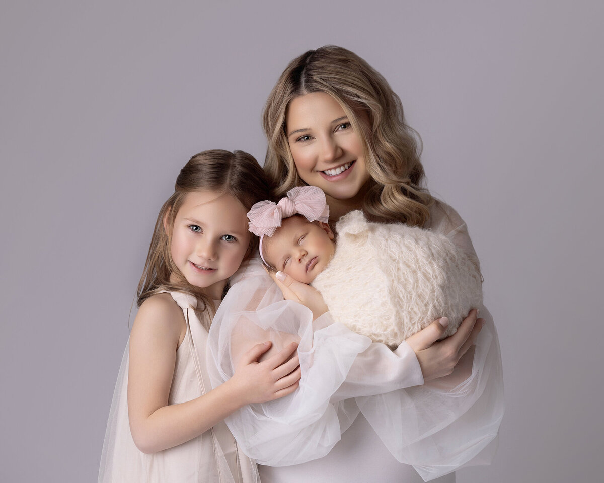 newborn session girl, mom is holding baby in her arms and her older daughter in leaning on her, hugging her mommy. Newborn baby girl is wrapped in creme crochet mohair wrap and is wearing a pink bow. Mom and older sister are dressed in white. Mommy and me session.