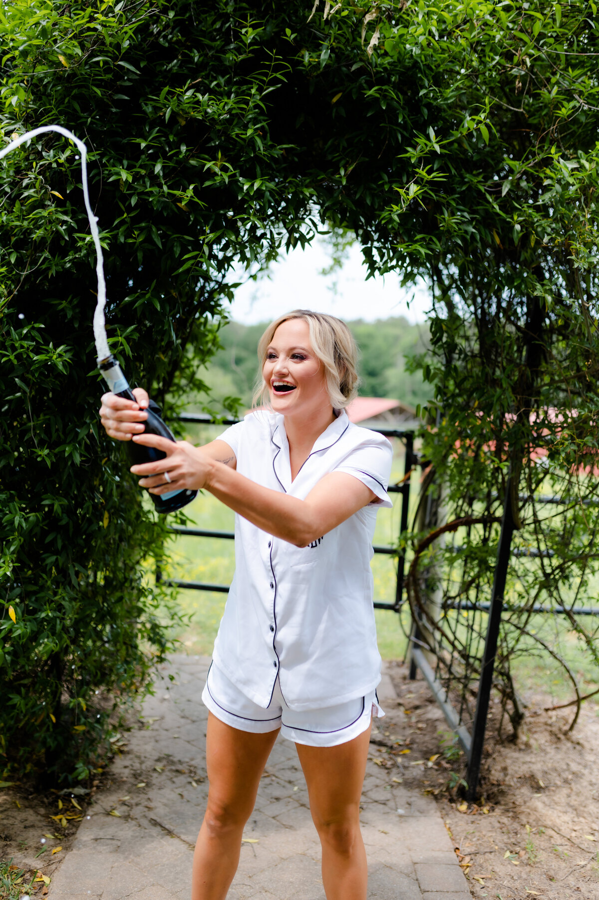 bride in a garden popping champagne the morning of her wedding day as she wears a white pajama set