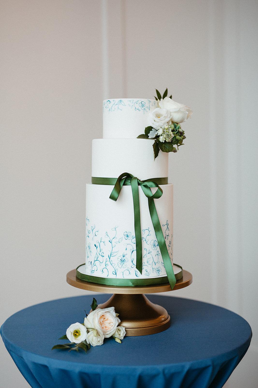 White cake with green ribbon and flowers on a gold cake stand atop a rounded table with blue linen.