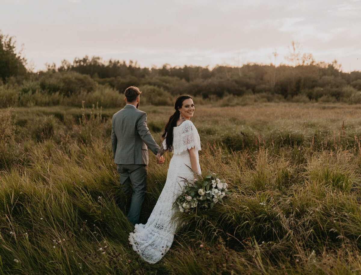 Bride and groom walking through field of tall grass, wearing a beautiful off-the-shoulder lace vintage boho lace gown, captured by Ash Maclean Photography, romantic elopement and wedding photographer in Red Deer, Alberta. Featured on the Bronte Bride Vendor Guide.