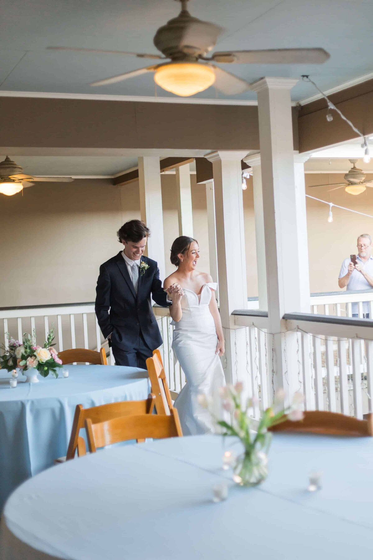 A bride in a white gown and a groom in a dark suit walk together down a balcony with white railings, round tables, wooden chairs, and floral centerpieces.