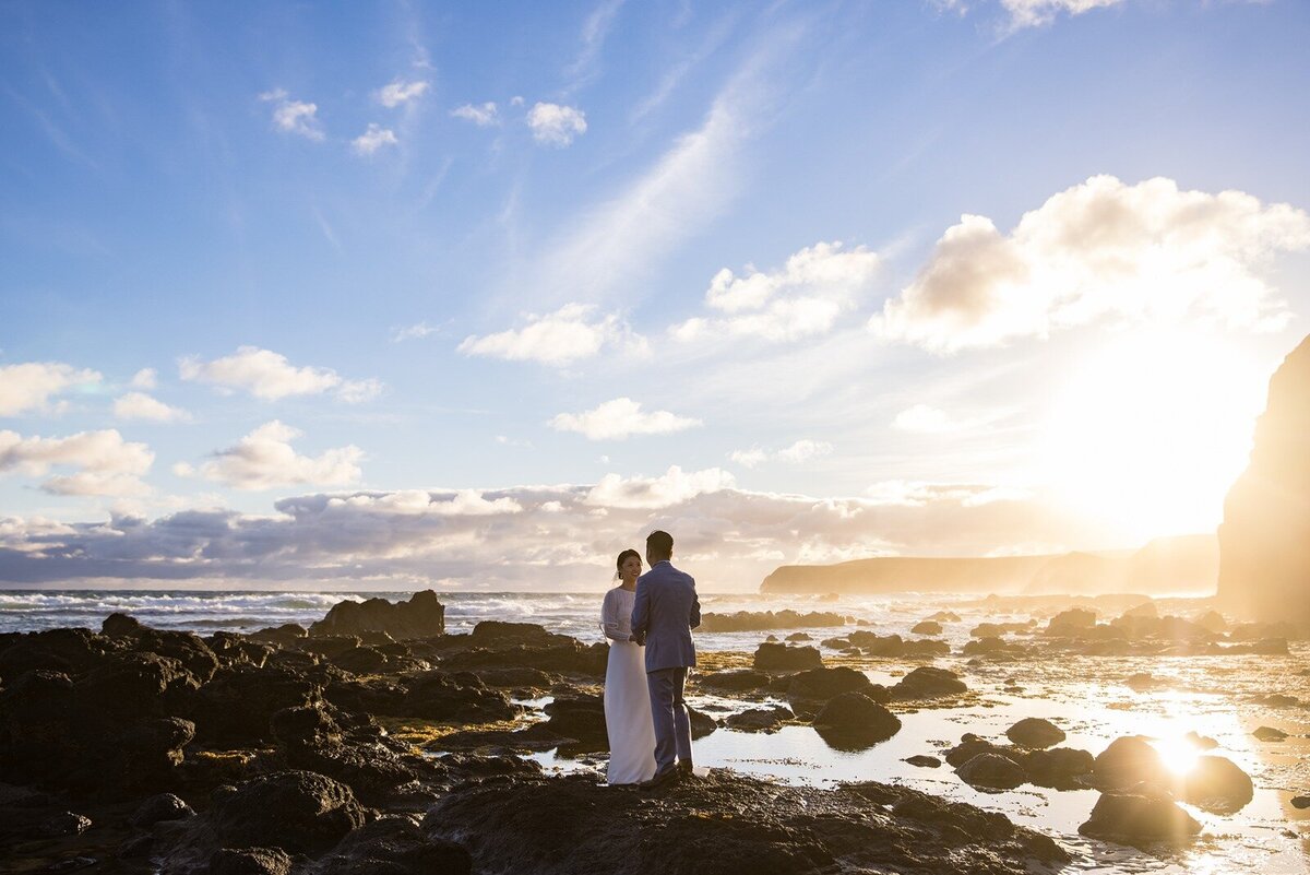 A bride and groom standing on rocks with the tide out and a beautiful sunset with scattered clouds
