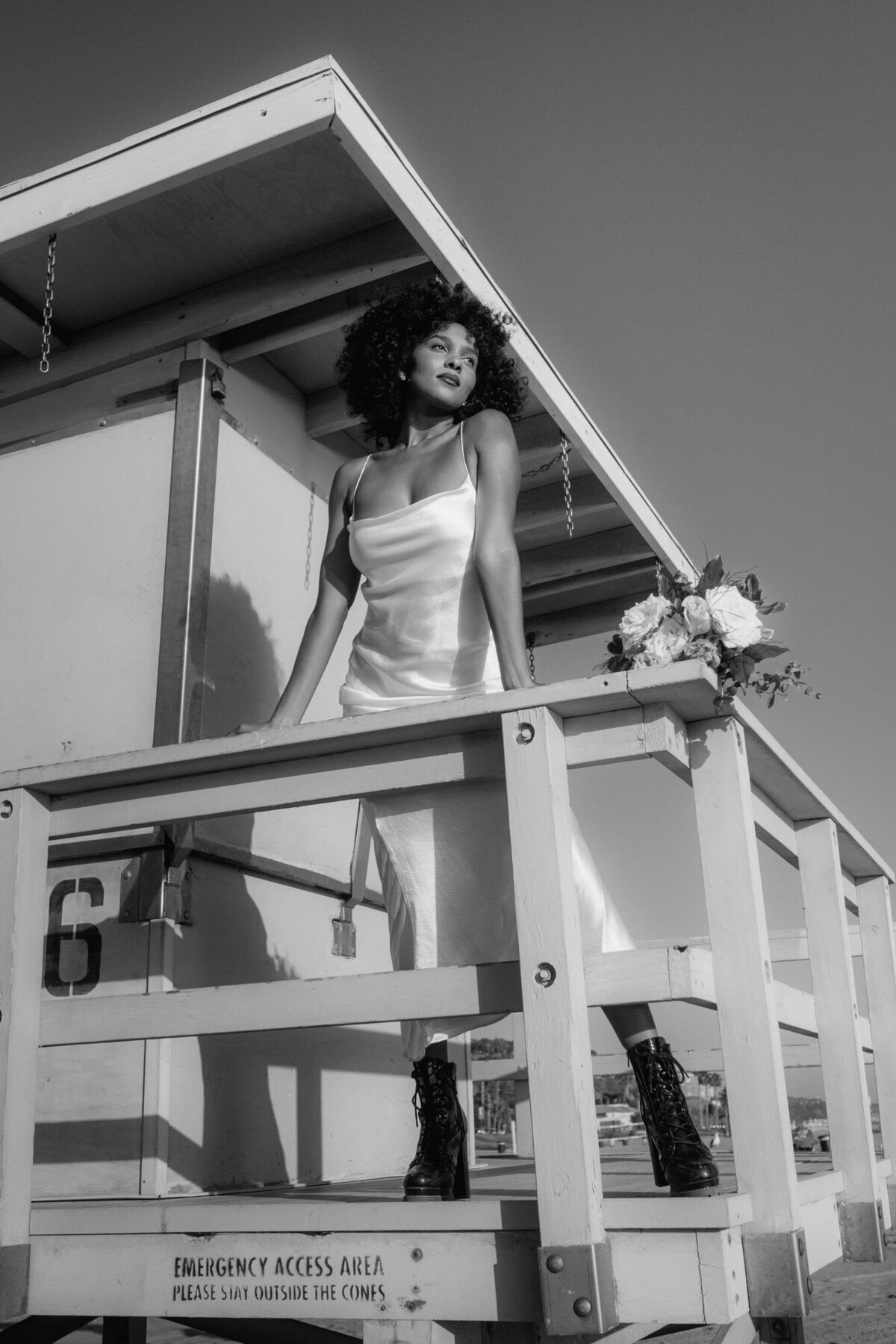 Im breitbeinigen Stand lehnt sich die Braut auf das Meer blickend an ein Geländer.