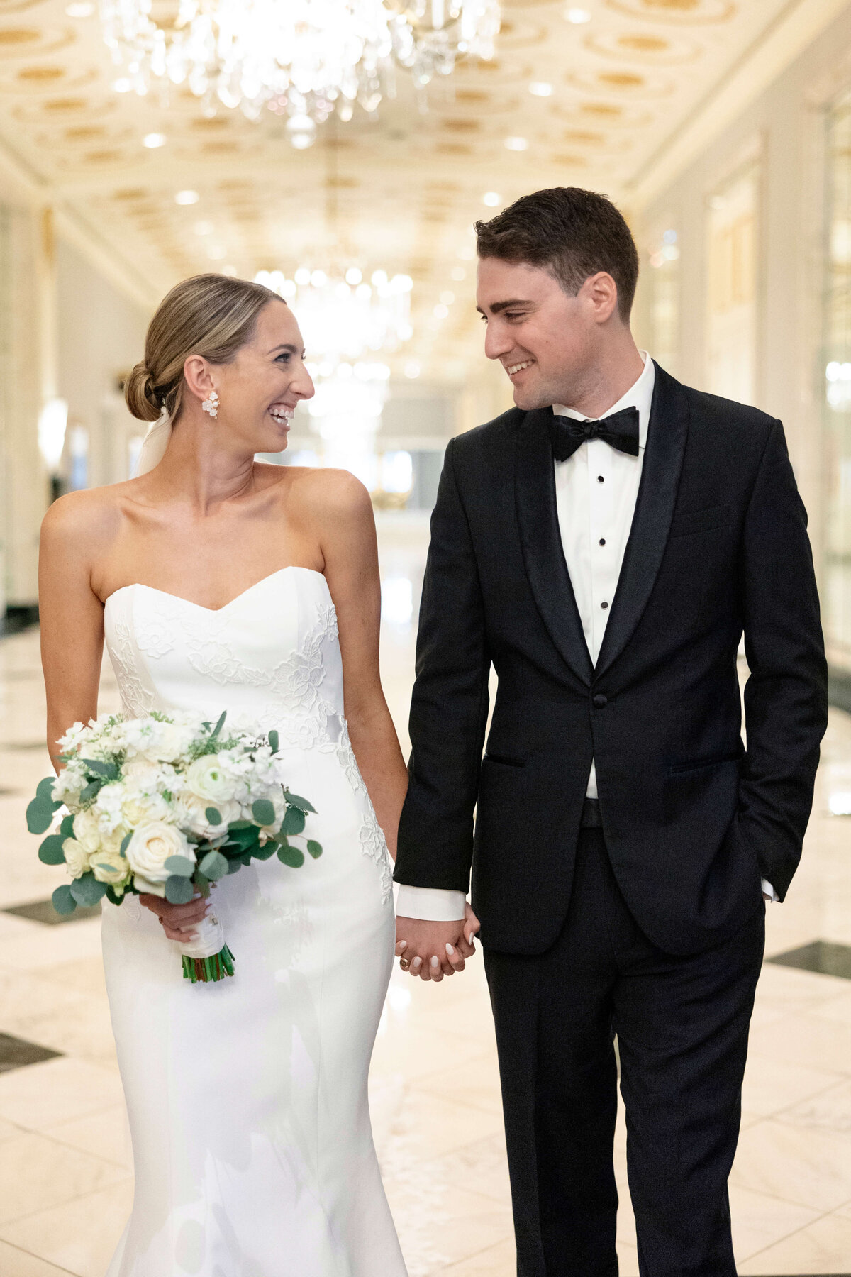 A bride in a white gown and a groom in a black tuxedo smile at each other while holding hands. She holds a bouquet of white flowers and greenery. They walk down a brightly lit hallway with chandeliers.