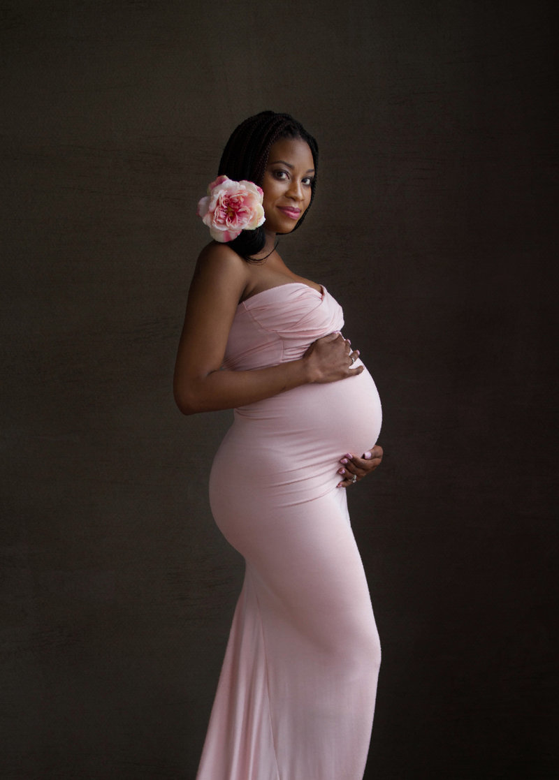 Beautiful African American woman maternity portrait against a grey hand-painted Vanity Fair style backdrop.  Woman is wearing a fitted pink gown and has a pink flower in her braided hair.