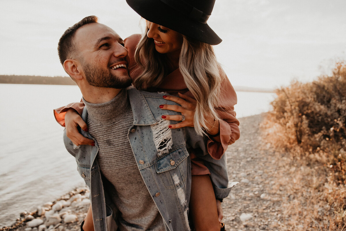 Gorgeous engaged couple, piggy back, laughing, captured by Kelsey Vera Photography, intimate and romantic wedding photographer in Airdrie, Alberta. Featured on the Bronte Bride Vendor Guide.