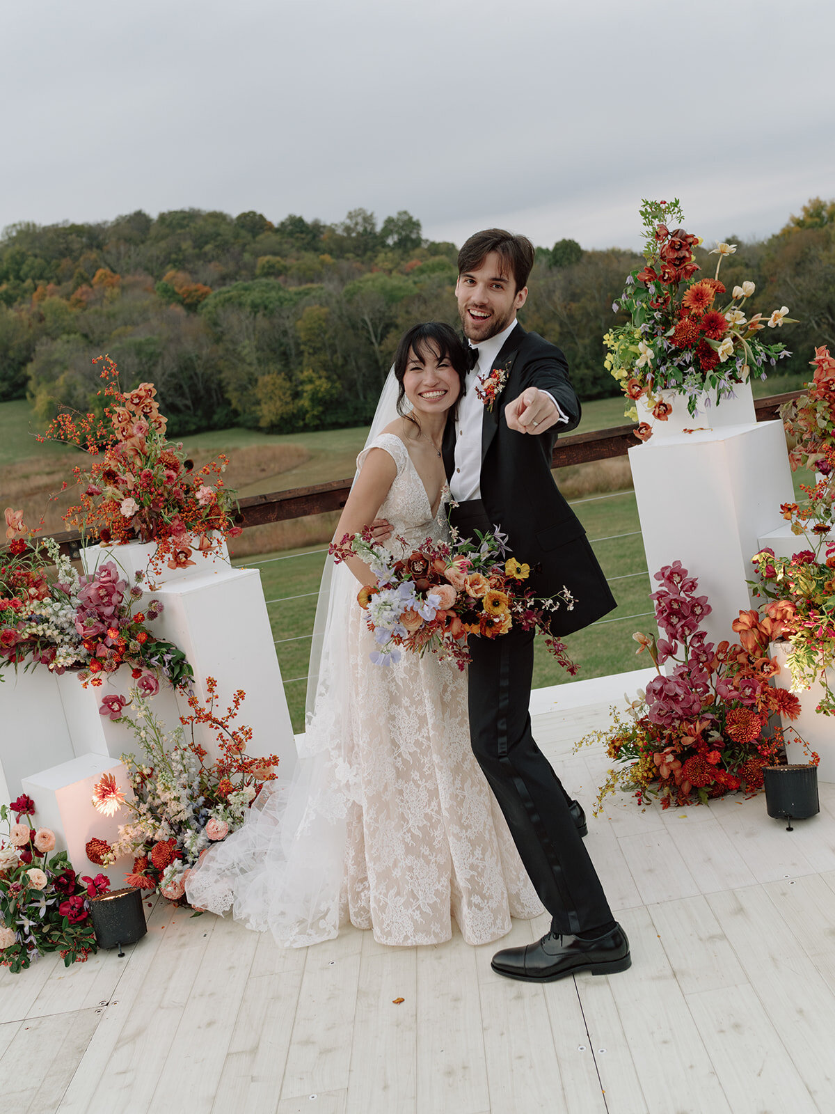 Sculptural columns wedding ceremony backdrop. Stair step white ceremony columns adorned with sculptural floral meadows in fall hues. Clear top tent build out for fall wedding reception in Tennessee hills. Wire threaded orchids, bittersweet berries, butterfly ranunculus, and tulips are highlighted in this wedding ceremony backdrop. Artistic fall color palette in copper, gold, ruby, lavender, and rust. Design by Rosemary & Finch Floral Design in Nashville, TN.