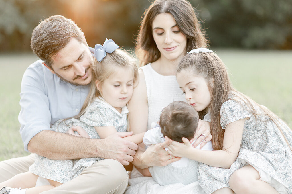 Big sisters admire their new baby brother as they sit with their parents at sunset