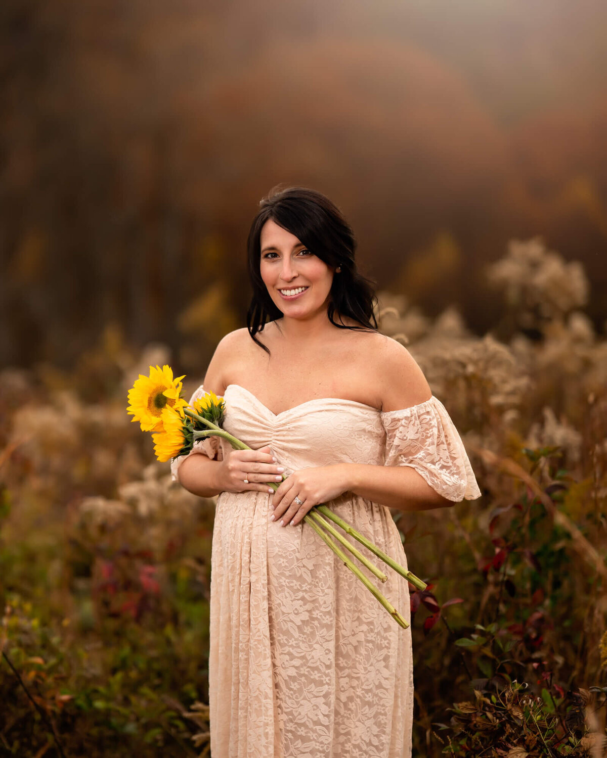 An expecting woman hold sunflowers across her belly while standing in front of tall grass
