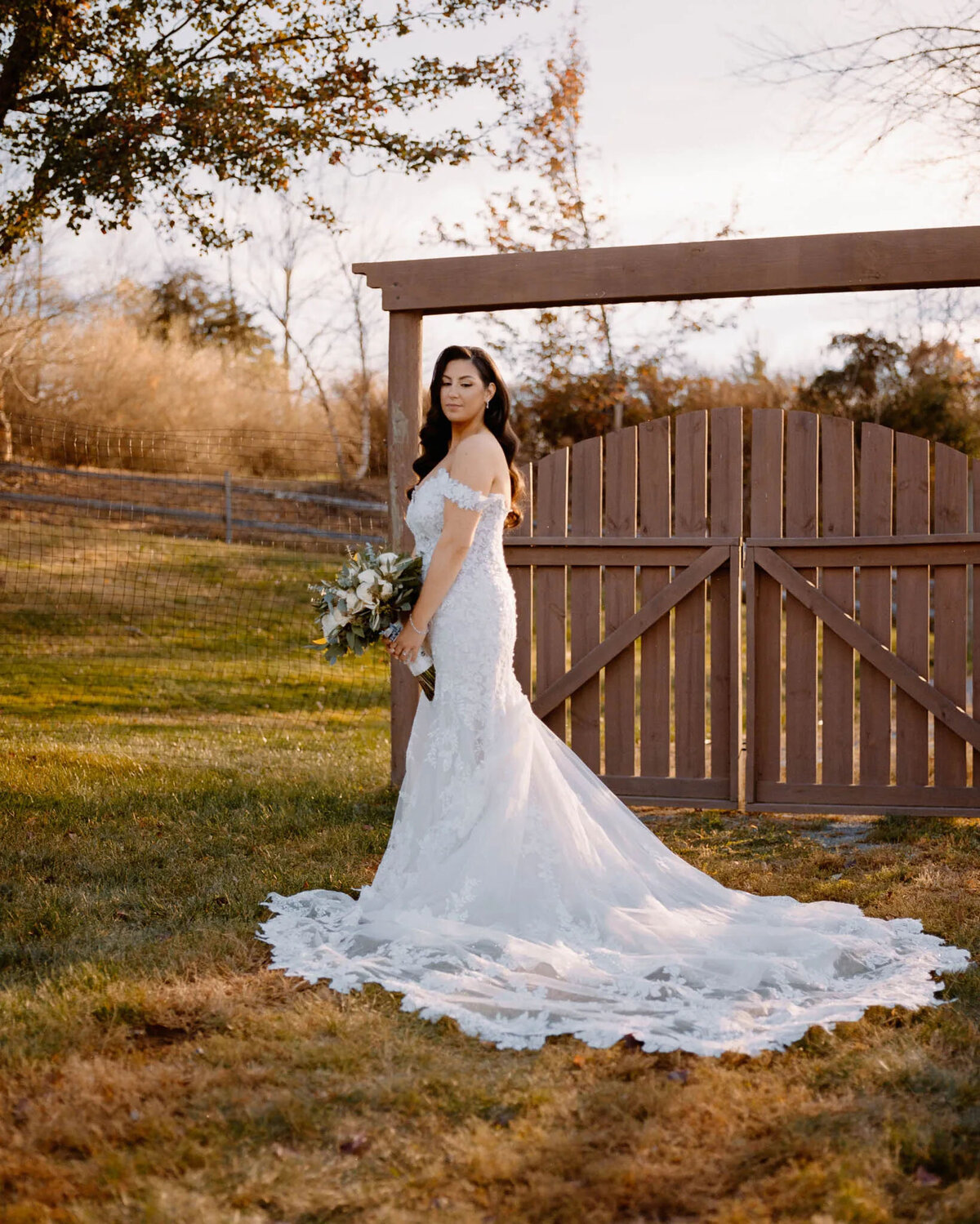 A bride in an off-shoulder lace wedding dress stands outdoors on grass, holding a bouquet, with a wooden gate and trees in the background, beautifully captured by an NJ wedding photographer. The dress features a long, elegant train spread out behind her.