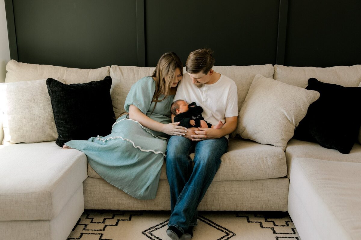 Young couple sitting on a cream-colored sectional couch, lovingly cradling their newborn baby in a cozy, natural light setting.