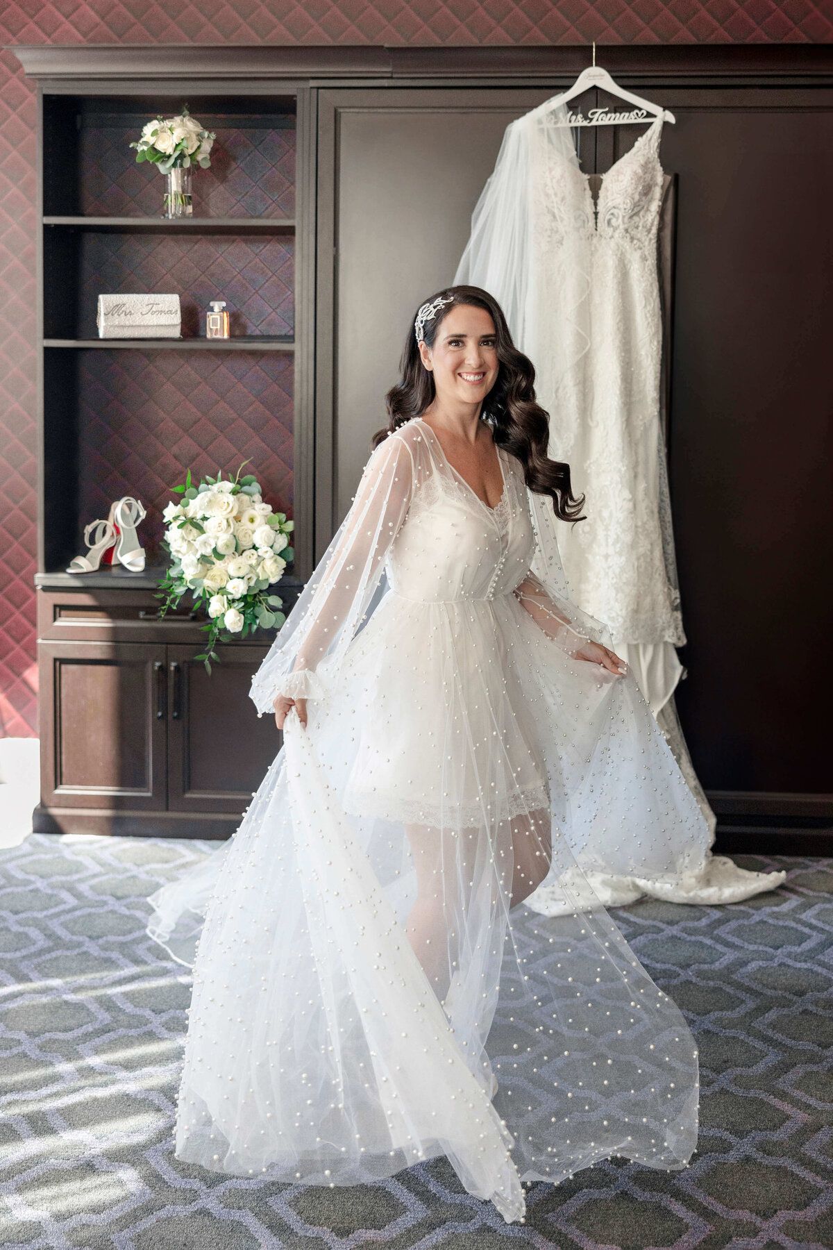 A bride smiles as she poses in a sheer, embellished robe in front of a hanging wedding dress. A bouquet and pair of shoes are displayed on a shelf behind her in the elegant room.