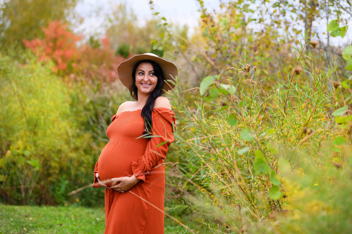 brown-haired pregnant lady wearing an orange dress and beige had standing in a field for her outdoor maternity photos in Burlington, On