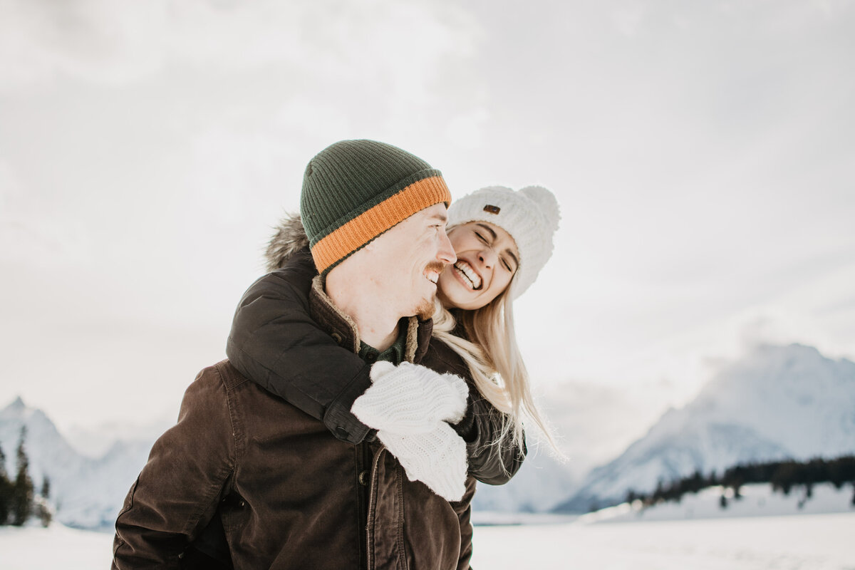 winter engagement session in the Grand Tetons with couple laughing and  smiling as the woman holds the man from around the neck