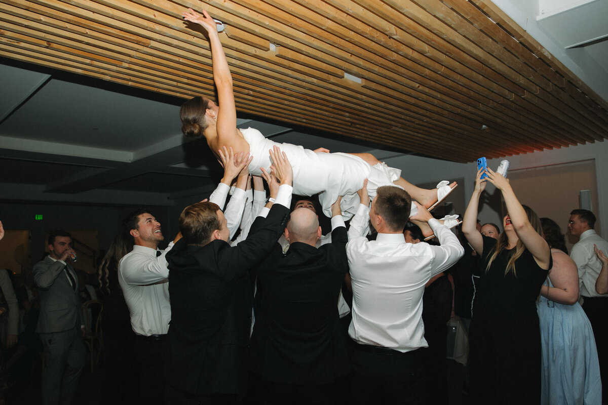 A bride in a white strapless, fitted wedding dress is held up in the air by several men in formal dress shirts and sports coat. She is horizontal and lifts her right hand into the air so that it almost touches the wood panel ceiling. 