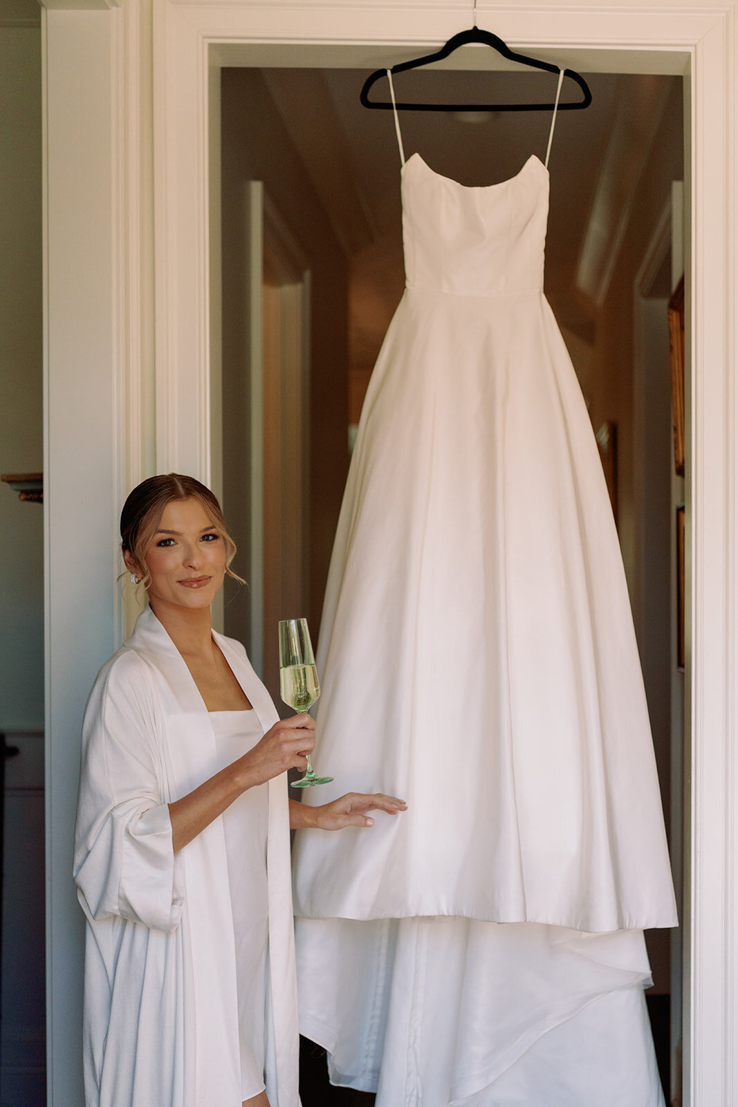 Bride holding a champagne glass and smiling beside her hanging wedding gown during getting ready at Burritt on the Mountain in Huntsville, Alabama, photographed by Perry Hancock.