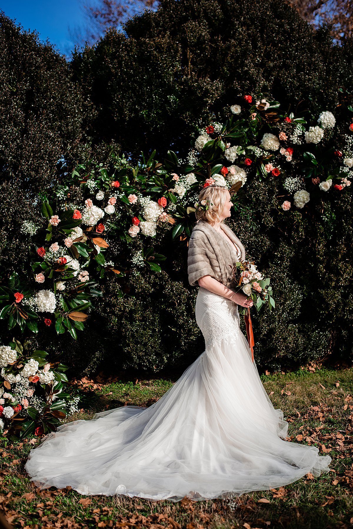 A bride in a light brown fur wrap with a flowing organza wedding dress with a fill train stands in front of a large floral installation of white hydrangea, magnolia leaves, blush roses and orange roses is a large boxwood hedge at Rippa Villa. The bride is holding a bouquet of blush and white flowers wrapped in a rust colored velvet ribbon which matches the flower crown in her hair for her winter wedding.