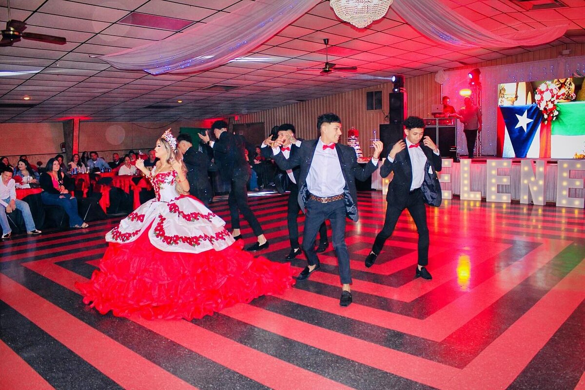 A Quinceañera wearing a big ball gown dancing next to chambelanes dressed in Quinceañera suits on a lit dance floor