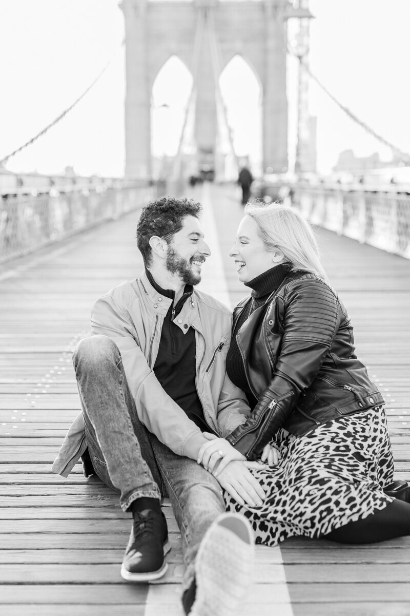 Sunset on the Brooklyn Bridge in NYC is the perfect place for an engagement session. This black and white photo is of an engaged couple sitting in the middle of this iconic bridge in New York City.