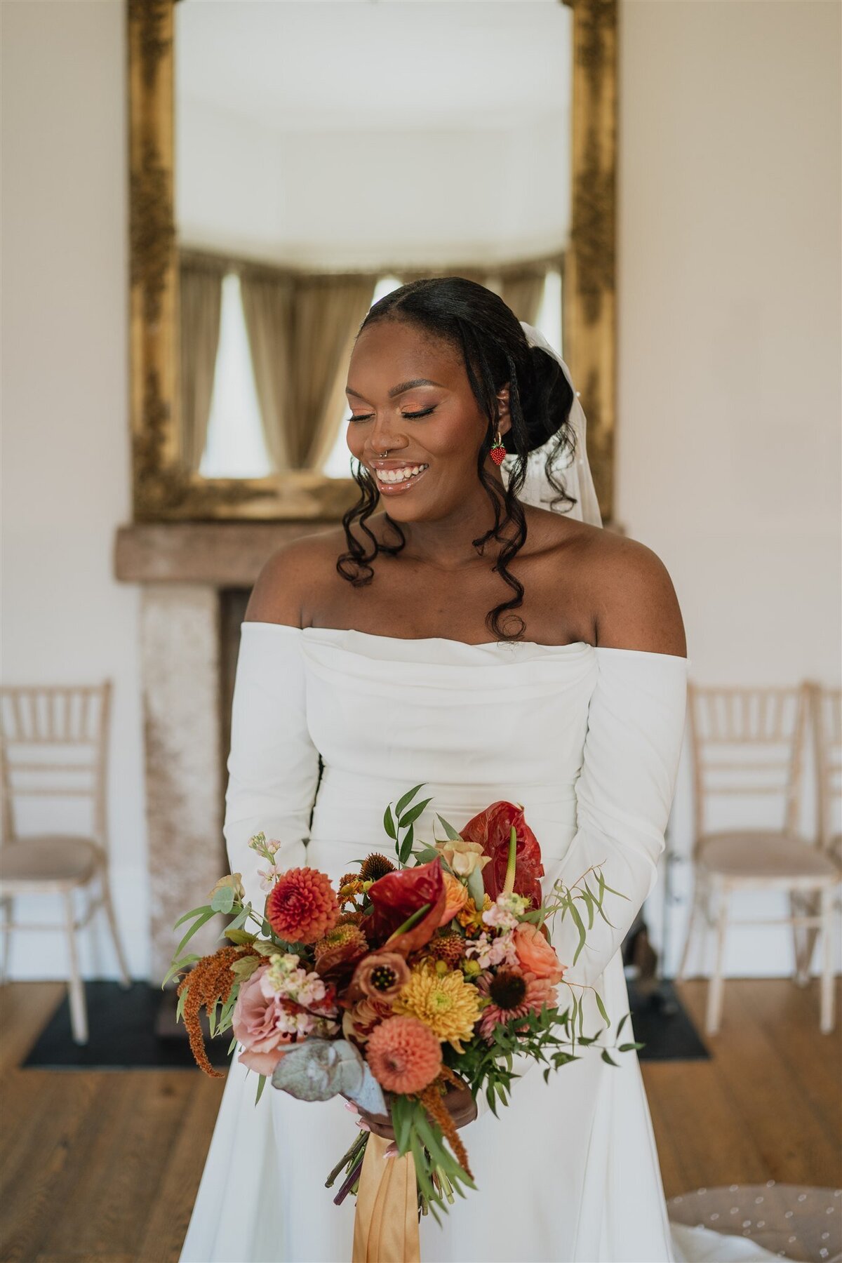 A bride wears a long white wedding dress and is photographed holding her bouquet in the empty dining room at Logie Country House. A large mirror hangs behind her and she smiles candidly.