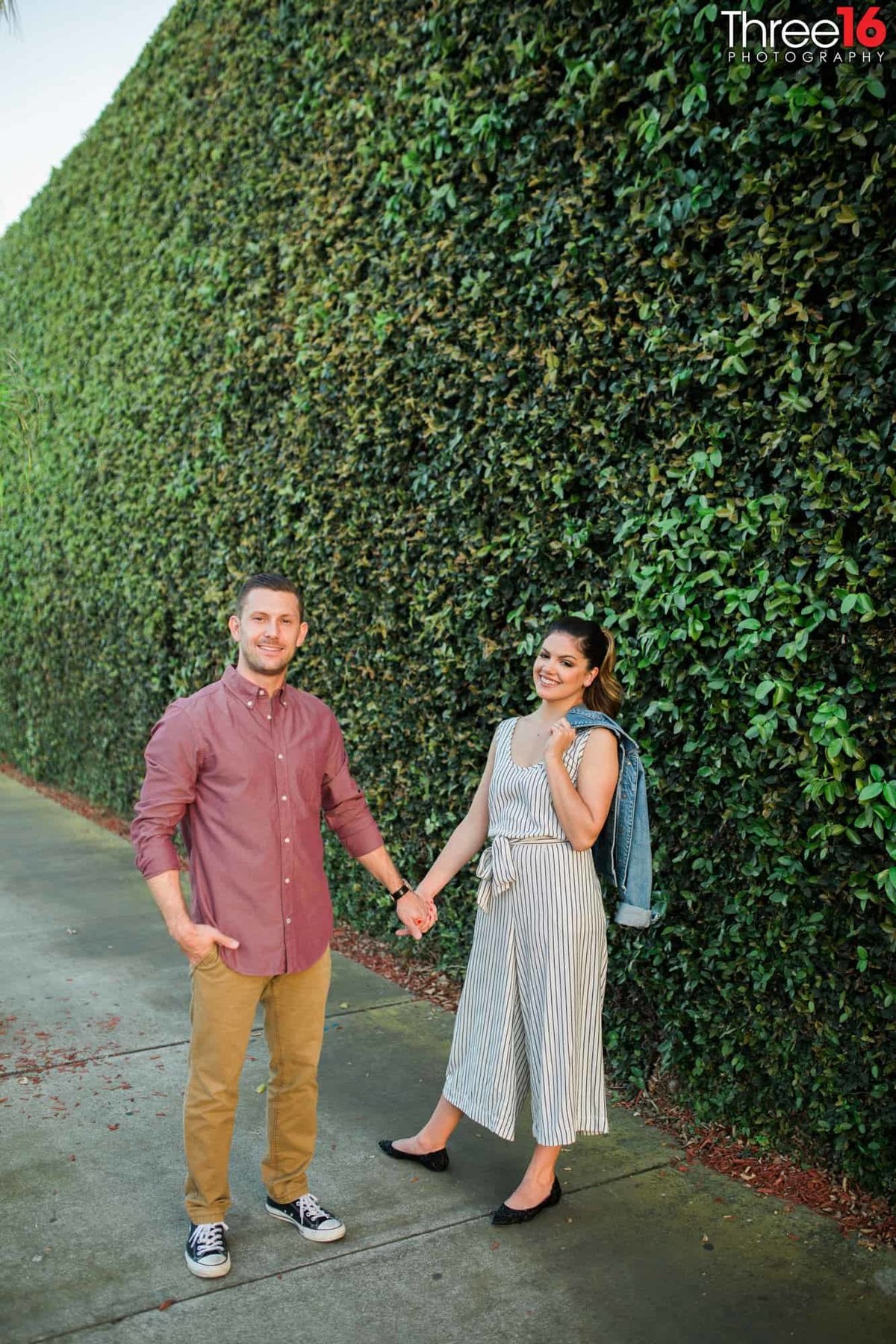 Engaged couple pose for photos while holding hands in front of a ivy-filled wall