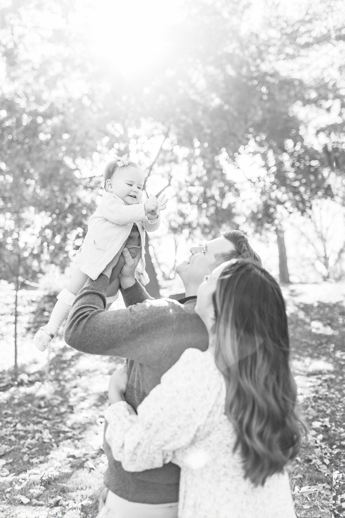 Black and white photo of father lifting his baby girl toward the sunlight during her first birthday session in a Kansas City park.