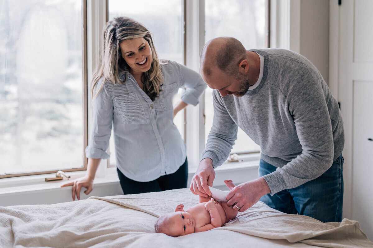 A father changes his new baby's diaper on the bed while his wife smiles, looking at at the baby.