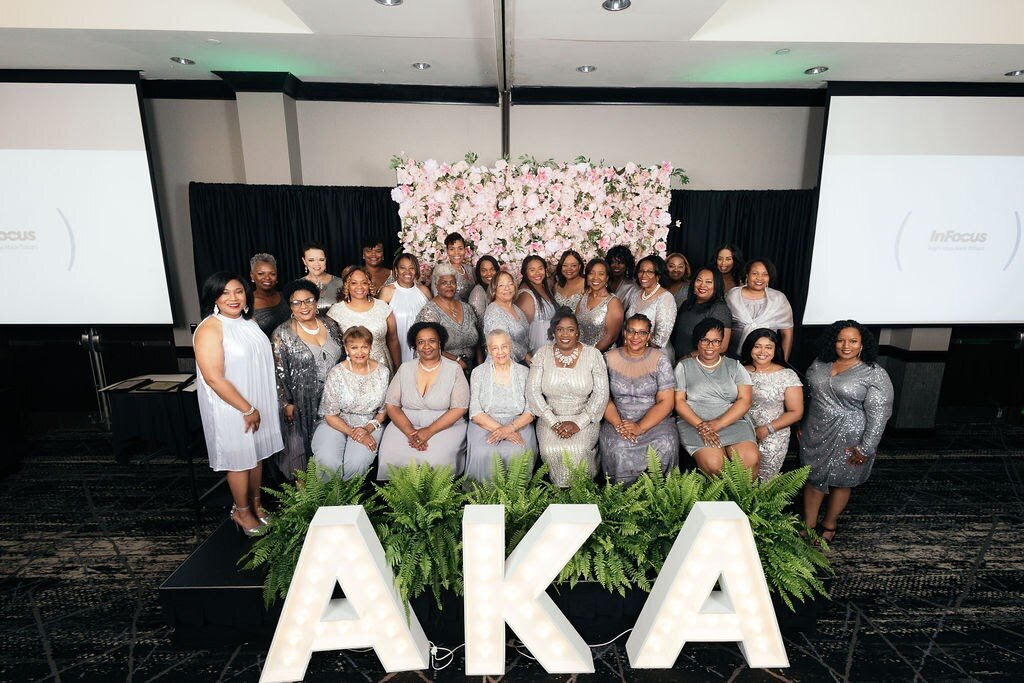 Phi Alpha Omega Charter Members wearing silver gowns in front of a flower wall and behind giant AKA letters
