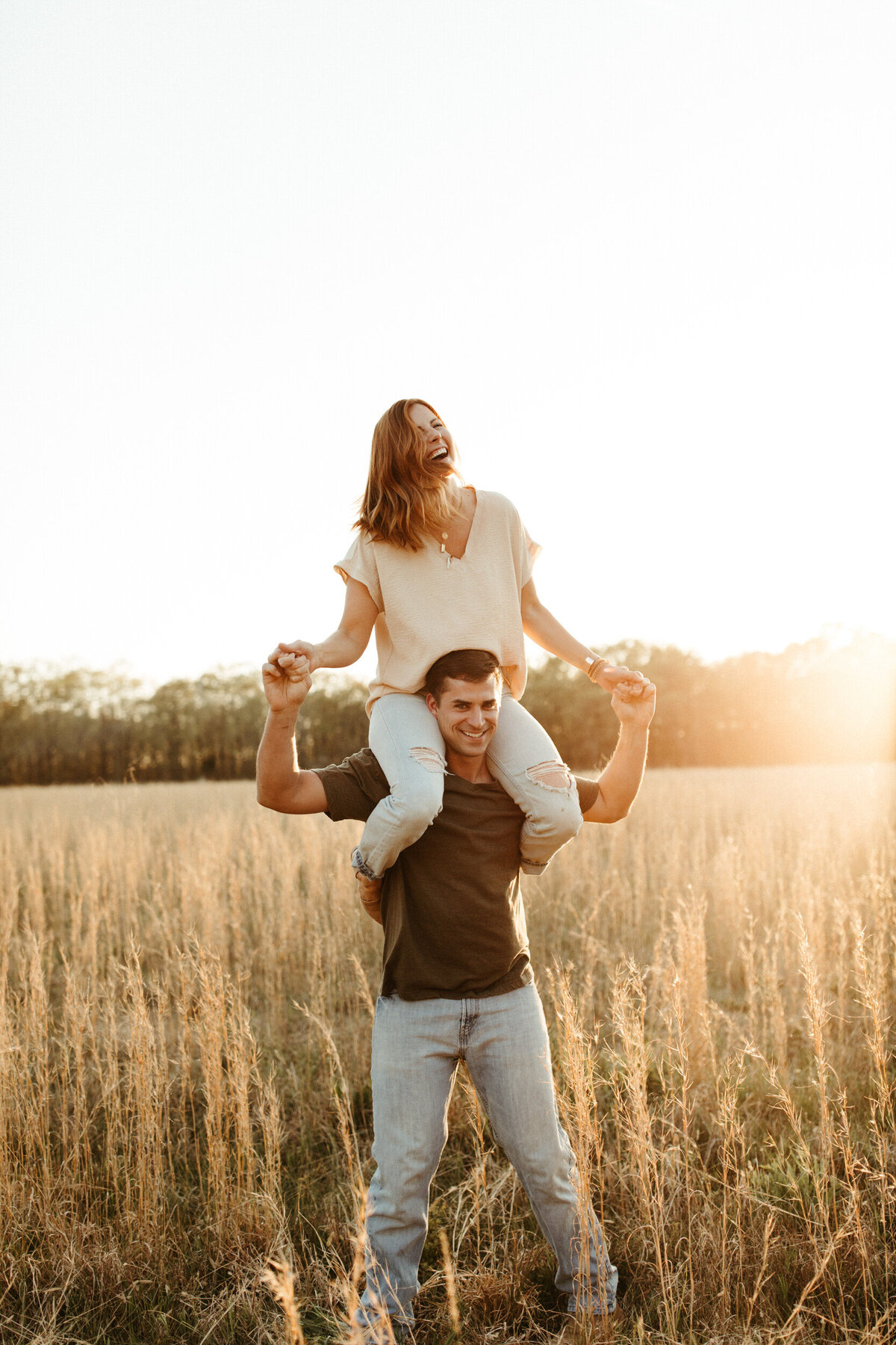 A girl is on top of a guy's shoulders as they laugh and walk through a field with tall grass at sunset.