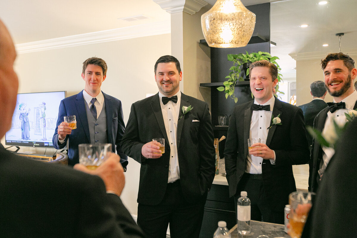 candid shot of the groom and groomsmen laughing during a toast at The Adolphus in Dallas, capturing joyful and lively moments before the wedding ceremony.
