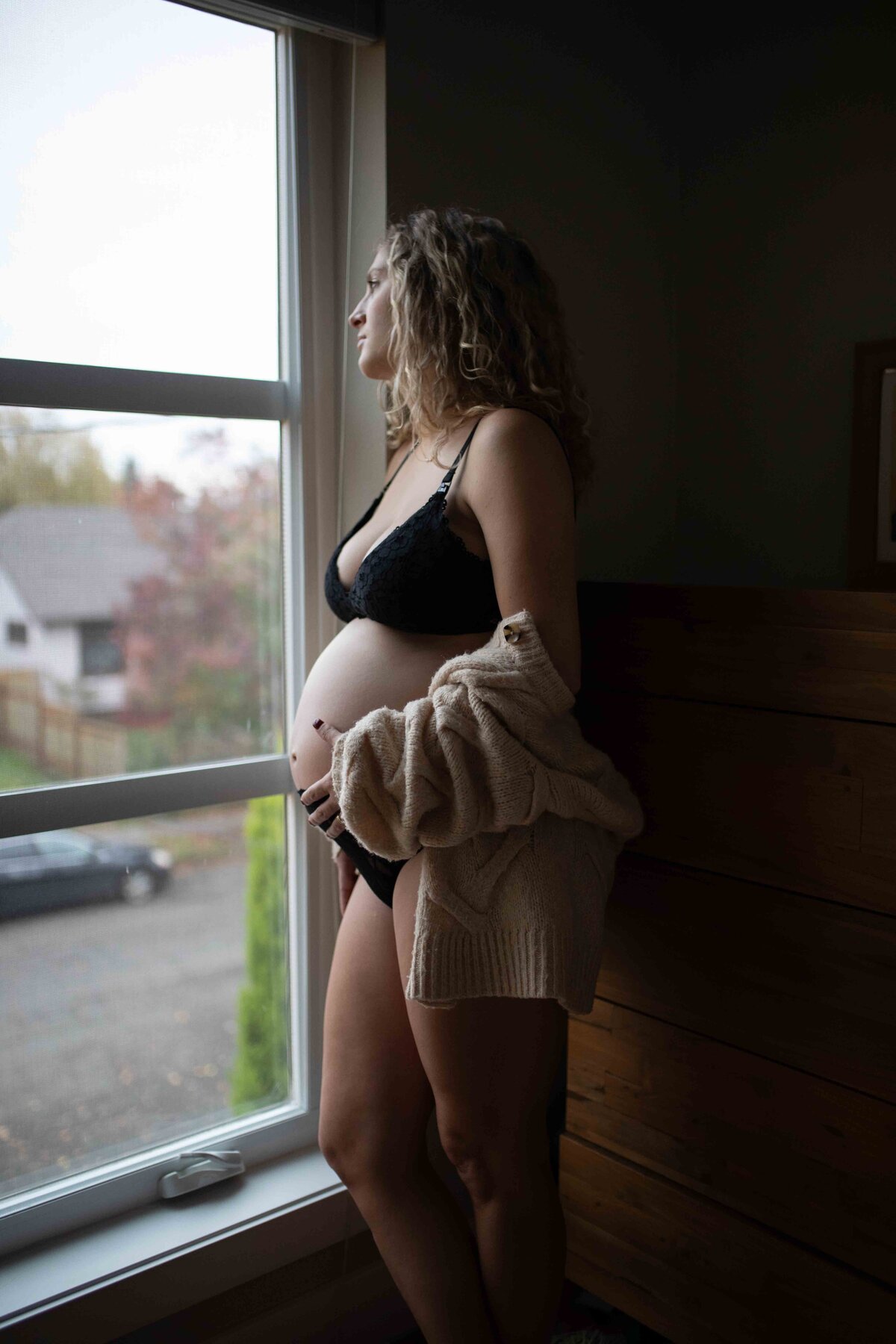 Soon to be mother holds her pregnant belly and gazes out the window during a home maternity photoshoot in Portland, Oregon.
