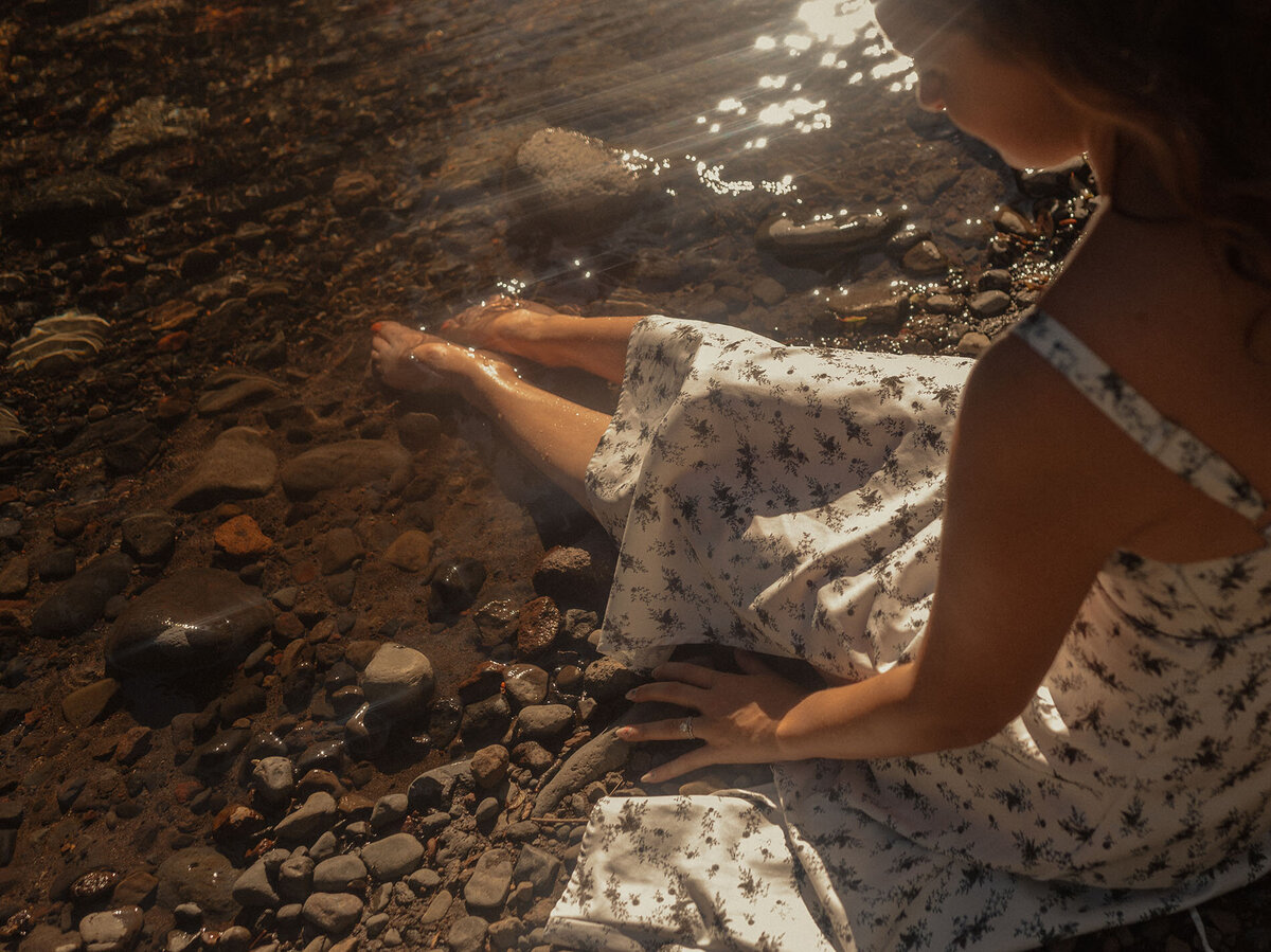 woman in white dress sits near the river and touches the water on a sunny warm day in Bend, Oregon
