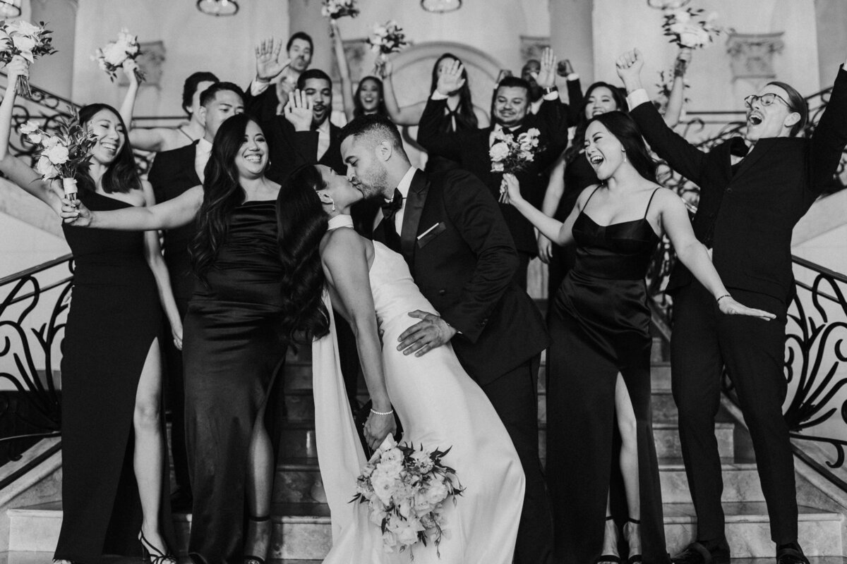 A couple leans over and kisses while their wedding party celebrates around them at The Stirling Club.