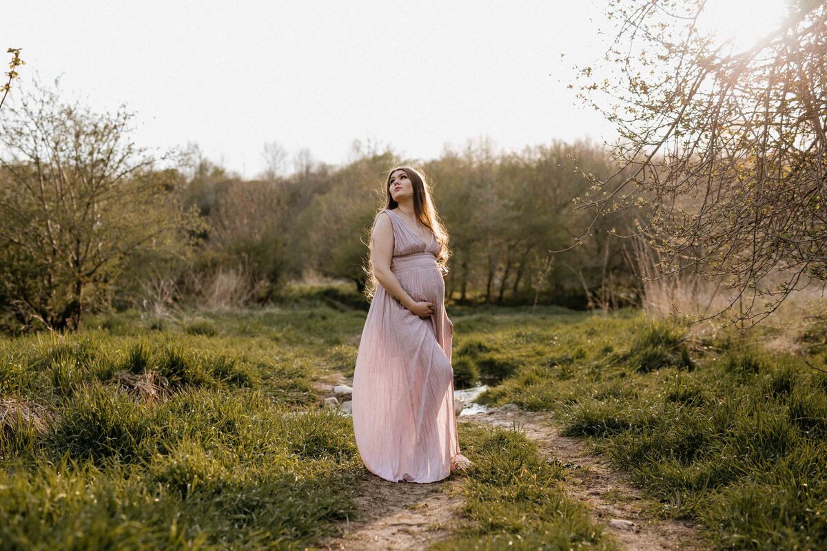 Maternity photo session in Exeter, Ontario field. Mom in long pink gown at holden hour. Her front hand is under her bump, her back hand off to her side. She is looking over her shoulder at the horizon.