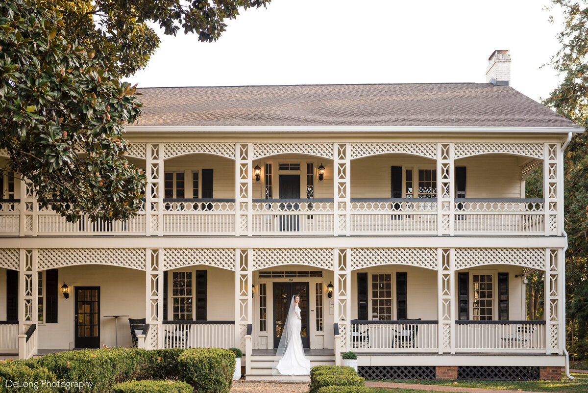 Wide angle outdoor bridal portrait taken on the front steps of the White Home in Rock Hill, SC. The entirety of the house in in the image. The bride is standing on the top step, her dress cascading down the steps below. She is looking over her right shoulder at the camera, smiling.
