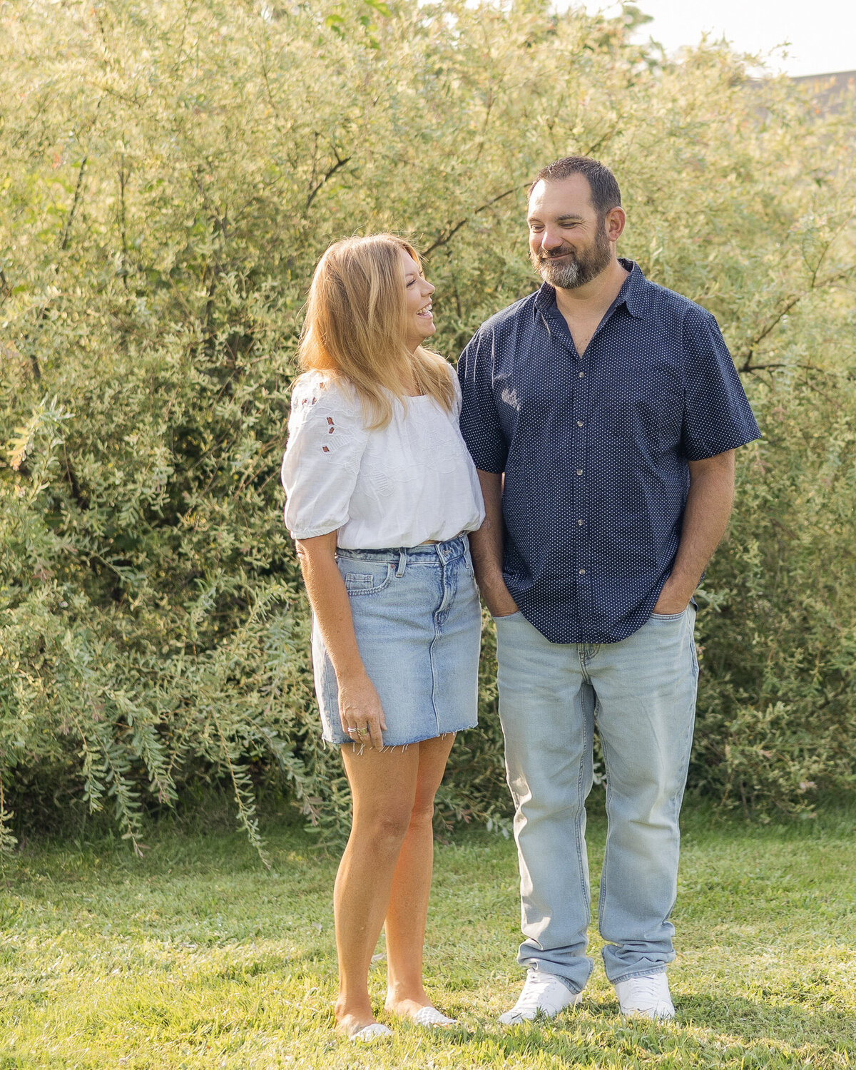 Husband and wife standing next to each other in grass with a bush behind them.  They are looking at each other smiling.  Wife is wearing a jean skirt, white half sleeve shirt, and has long blonde hair.  Husband is wearing a blue button up short sleeve shirt, blue jeans, and has black gray short hair and beard.