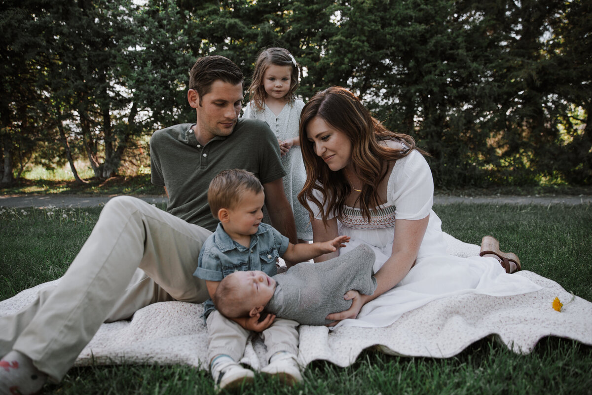 Idaho Falls newborn photos with mother father, and their three children sitting on a white blanket together for their summer newborn photos as the entire family looks at the newborn