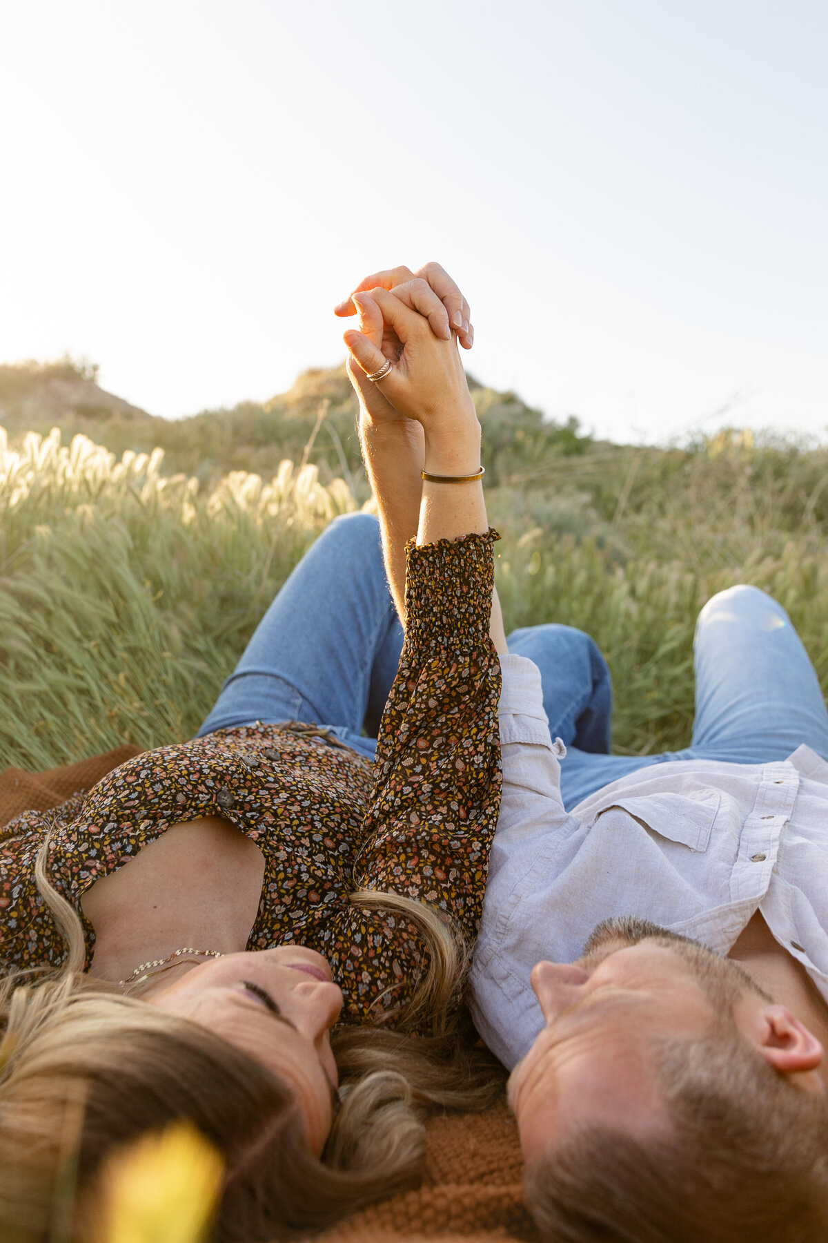 A couple lies in a grassy field in Saratoga Springs, Utah, holding hands against the backdrop of the sky. The woman’s engagement ring shines as they intertwine their fingers.