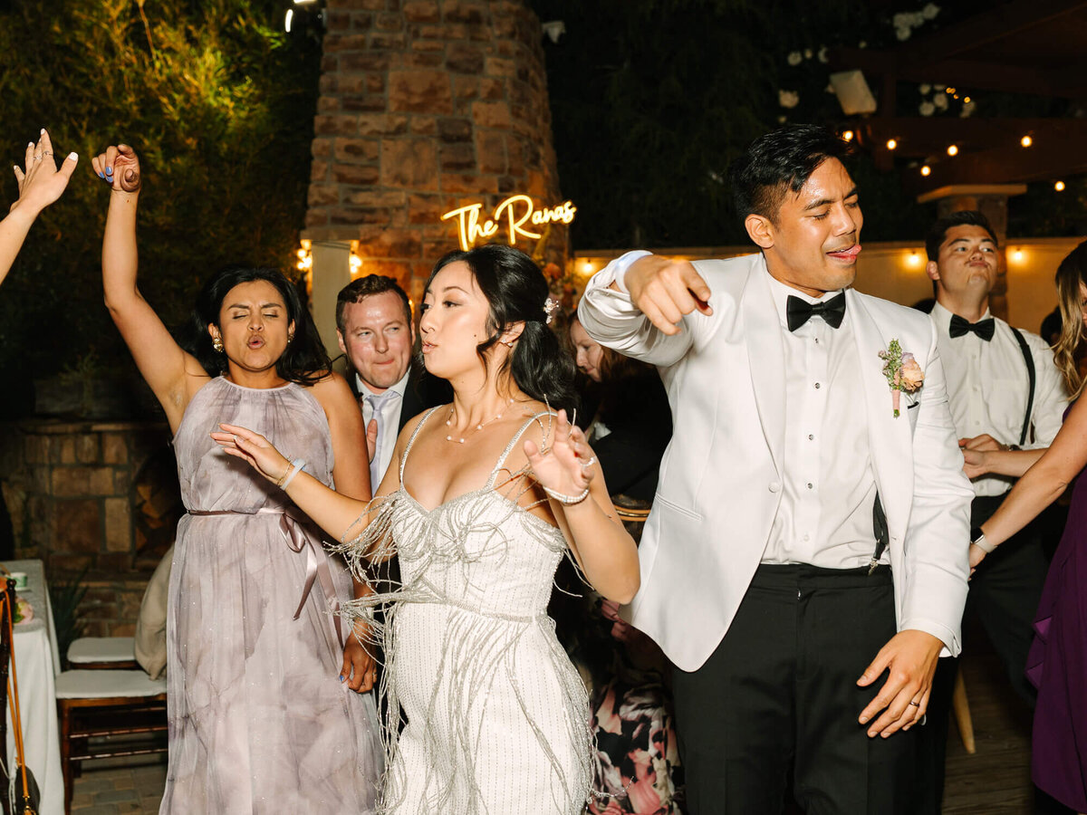 Wedding guests dance energetically in formal attire, with joyful expressions. A neon sign reads "The Ranas" in the background.