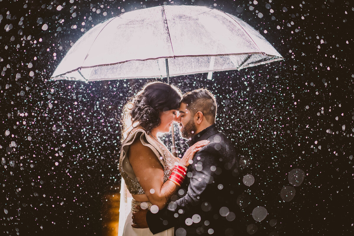 Indian bride and groom under an umbrella on a rainy evening lit up by a flash behind them